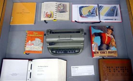 left display case, showing an olh brailler and several books in braille and print-braille.